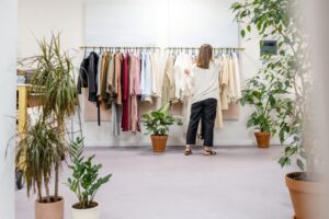  Kvinde ser på tøj på et stativ i en moderne butik med lyse vægge, farverigt tøj og grønne planter i potter.